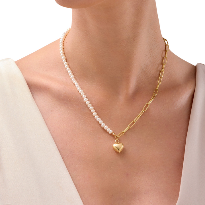 A person wearing an 18K gold necklace with a paperclip chain, freshwater pearl strand, and an 18K gold love heart pendant.